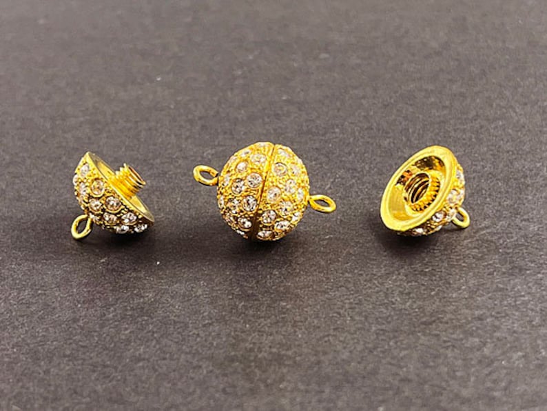 Op de afbeelding: Drie goudkleurige metalen sluitingen met heldere kristallen versieringen. Elke sluiting heeft een schroefontwerp en een lus om aan een ketting of armband te bevestigen.