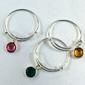 May include: Three silver rings with dangling charms. The charms are round and feature different colored gemstones: pink, green, and amber.