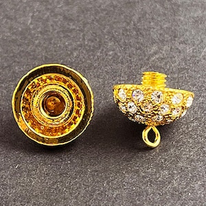Op de afbeelding: Een gouden metalen dop met een helder kristalontwerp. De dop heeft een kleine lus aan de onderkant om aan een ketting of armband te bevestigen.