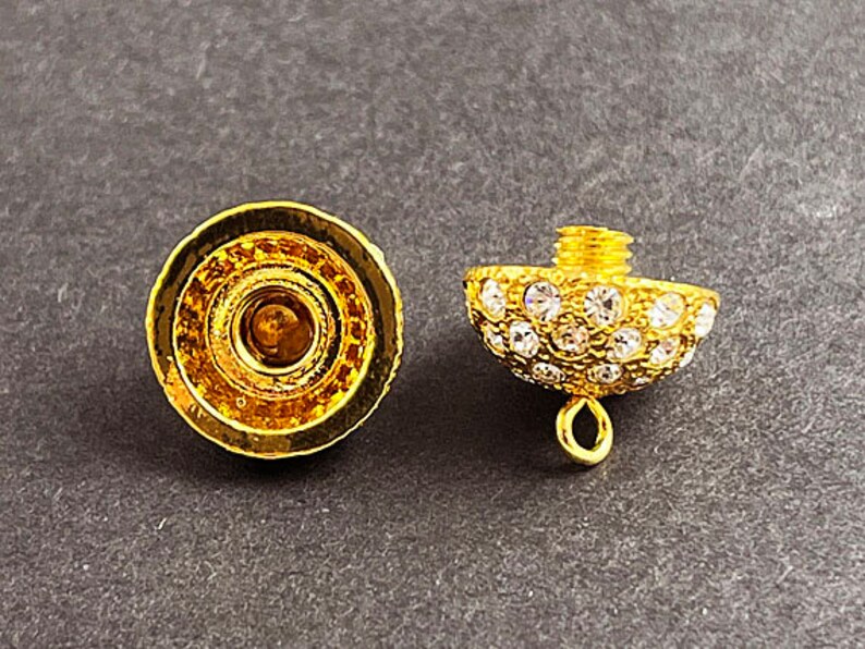 Op de afbeelding: Een gouden metalen dop met een helder kristalontwerp. De dop heeft een kleine lus aan de onderkant om aan een ketting of armband te bevestigen.
