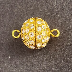 Op de afbeelding: Gouden magnetische sluiting met heldere ronde strass steentjes. De sluiting is rond en heeft een gladde, gepolijste afwerking.