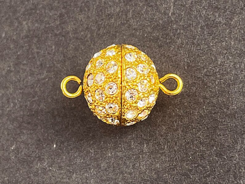 Op de afbeelding: Gouden magnetische sluiting met heldere ronde strass steentjes. De sluiting is rond en heeft een gladde, gepolijste afwerking.