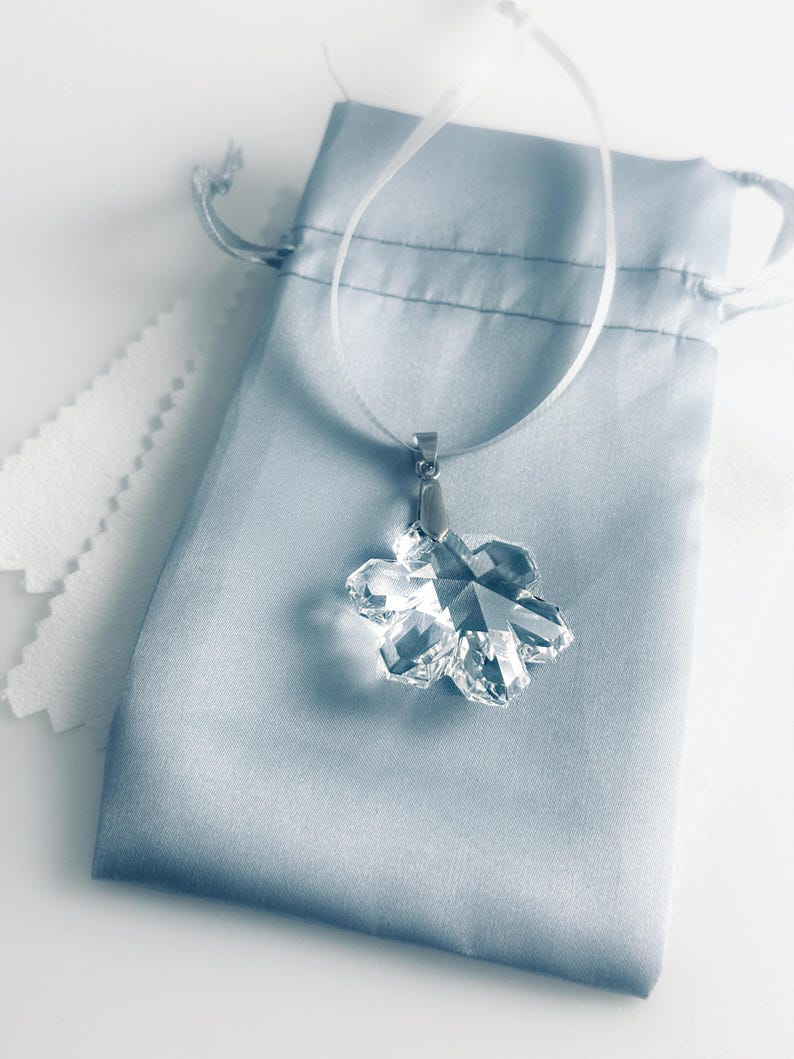 Swarovski Magic Classic Crystal snowflake ornament .A silver snowflake pendant necklace with a clear crystal snowflake charm. The necklace has a white satin ribbon.