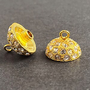Op de afbeelding: Twee gouden metalen kralencaps met heldere strasssteentjes. De caps zijn rond en hebben een klein lusje aan de bovenkant om aan sieraden te bevestigen.