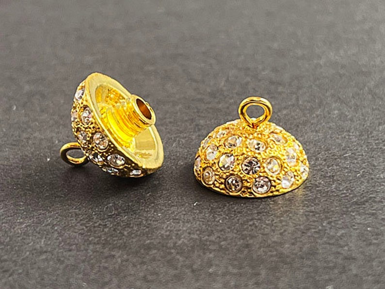 Op de afbeelding: Twee gouden metalen kralencaps met heldere strasssteentjes. De caps zijn rond en hebben een klein lusje aan de bovenkant om aan sieraden te bevestigen.