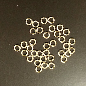 May include: A group of gold jump rings, scattered on a black surface. These rings are used for jewelry making and crafting.
