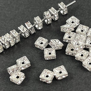 May include: A collection of silver-toned, square-shaped, rhinestone-encrusted embellishments. The embellishments are arranged in a variety of patterns and sizes.