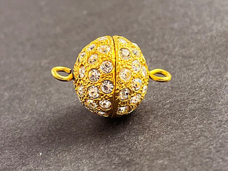 Op de afbeelding: Gouden magnetische sluiting met heldere strass steentjes. De sluiting is rond en heeft aan beide zijden twee lussen.
