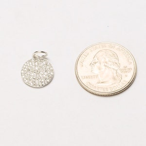 May include: A silver charm with a round, pave-set diamond design. The charm is approximately the size of a US quarter.