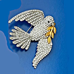 May include: A silver-toned dove brooch encrusted with clear crystals. The dove holds a gold-colored olive branch in its beak. The brooch has a pin on the back for secure attachment. The wings are spread in flight, and the brooch is set against a blue background.