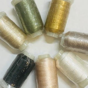 May include: Eight spools of thread in various colors, including green, gold, beige, black, and white. The spools are arranged in a circle on a white surface.