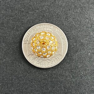 Op de afbeelding: Een gouden knop met een rond, verhoogd ontwerp. De knop is bedekt met kleine, heldere strass steentjes. De knop wordt getoond naast een Nederlandse 1 euro munt voor groottevergelijking.