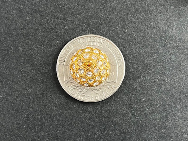 Op de afbeelding: Een gouden knop met een rond, verhoogd ontwerp. De knop is bedekt met kleine, heldere strass steentjes. De knop wordt getoond naast een Nederlandse 1 euro munt voor groottevergelijking.