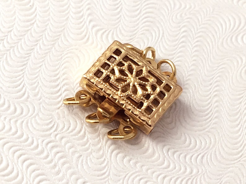 May include: Gold-toned metal clasp with a decorative filigree design. The clasp has multiple loops for attaching jewelry.