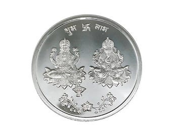 May include: A silver coin featuring two seated figures, possibly deities, on lotus flowers. The coin has embossed details and text at the top. A floral design is at the bottom. The coin is round with a shiny, reflective surface.