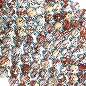 Op de afbeelding: Close-up van een kralenketting met ronde, patroonvormige kralen in tinten bruin, cr&egrave;me en wit. De kralen zijn geregen op lichtblauw koord, waardoor een textuur ontstaat. De kralen hebben een unieke, gespikkelde uitstraling.