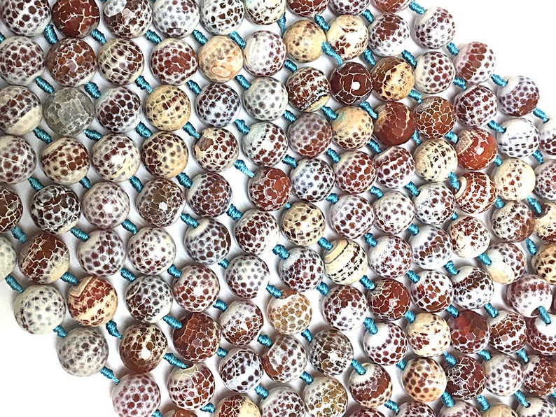 Op de afbeelding: Close-up van een kralenketting met ronde, patroonvormige kralen in tinten bruin, cr&egrave;me en wit. De kralen zijn geregen op lichtblauw koord, waardoor een textuur ontstaat. De kralen hebben een unieke, gespikkelde uitstraling.
