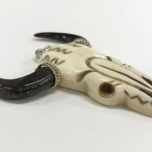 Peut inclure: Un pendentif en os de cr&acirc;ne de taureau blanc avec des accents de cornes noires et un enroulement de fil d'argent. Le cr&acirc;ne est sculpt&eacute; avec des d&eacute;tails complexes et a un trou en haut pour l'accrocher.