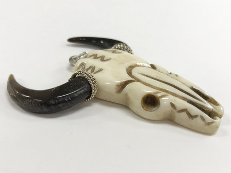 Peut inclure: Un pendentif en os de cr&acirc;ne de taureau blanc avec des accents de cornes noires et un enroulement de fil d'argent. Le cr&acirc;ne est sculpt&eacute; avec des d&eacute;tails complexes et a un trou en haut pour l'accrocher.