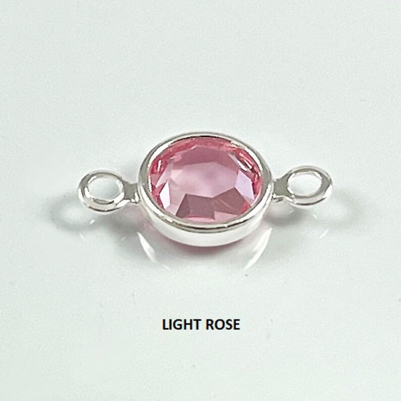 Puede incluir: Un conector plateado con un cristal de color rosa claro en el centro. El conector tiene dos bucles a cada lado.