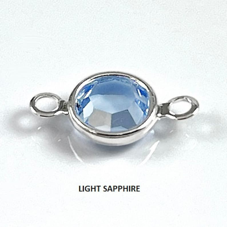 Puede incluir: Un conector plateado con un cristal azul claro en el centro. El conector tiene dos bucles a cada lado.