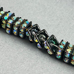 May include: A black hair clip with iridescent rhinestones set in a square pattern. The clip is made of metal and has a spring-loaded mechanism.