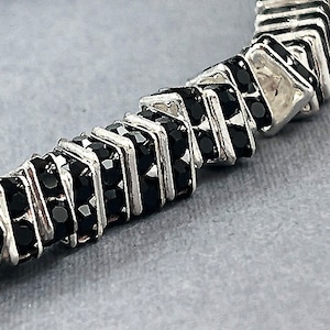 May include: A silver and black bracelet with a geometric design. The bracelet is made of small, square-shaped beads that are alternating silver and black.