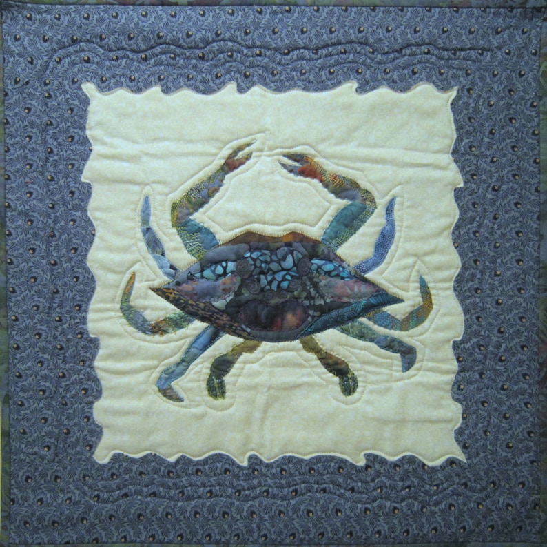 May include: A blue and brown quilted crab applique on a cream background with a blue and gray border. The crab is surrounded by a wavy border.