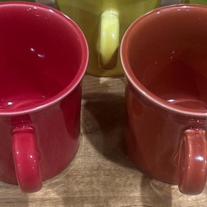 May include: Close-up of several ceramic mugs in various colors. The mugs have a rounded shape with a handle on the side. The mugs are red, orange, and green. The mugs are sitting on a wooden surface.