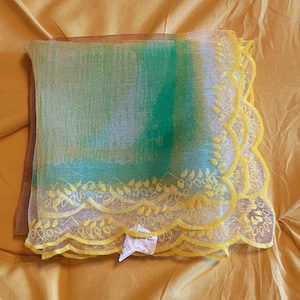 May include: A square sheer scarf featuring a gradient of green and white tones. The edges are finished with a scalloped border in bright yellow, decorated with an intricate embroidered leaf pattern. A small white fabric tag is attached to the corner.