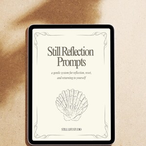Op de afbeelding: Een zwarte tablet toont een crèmekleurige pagina met de tekst "Still Reflection Prompts" en een schelpenillustratie. De tekst hieronder luidt "a gentle system for reflection, reset, and returning to yourself". De tablet ligt op een lichtbruin oppervlak.