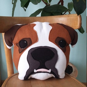 Könnte beinhalten: Ein braunes und weißes Bulldoggen-Gesichtskissen. Das Kissen ist aus Filz und hat eine angenähte Nase und Augen. Das Kissen liegt auf einem Holzstuhl.