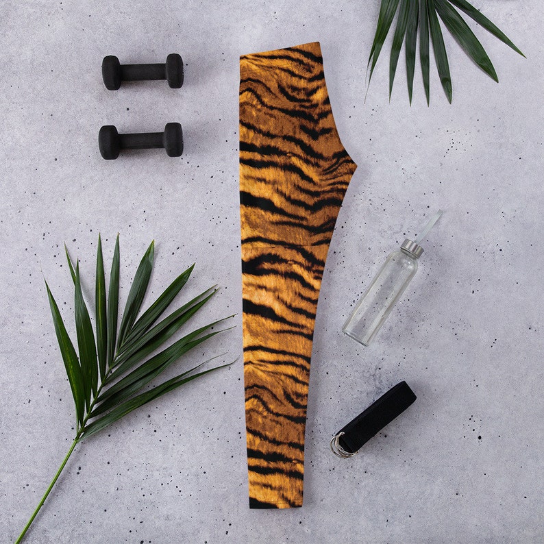 May include: A pair of black and orange tiger print leggings with a black yoga strap, a clear water bottle, and two black dumbbells on a gray concrete background.