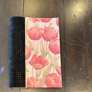 Op de afbeelding: Een hardcover notitieboek met een zwart-witte pied-de-poule rug en een omslag met een bloemen tulpenontwerp in roze, rood en groen. Het notitieboek is gesloten en ligt op een houten oppervlak.
