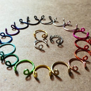 May include: A set of 18 colorful wire nose rings arranged in a circle on a brown surface. The rings are made of different colors, including purple, blue, green, gold, and red.