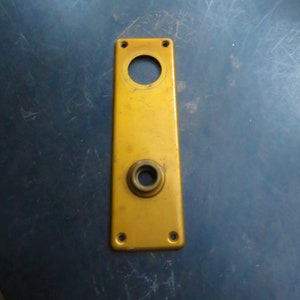 May include: A gold-colored metal door plate with a round hole and a smaller round hole with a metal knob. The plate has four small holes around the edges.