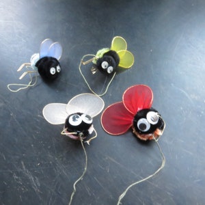 May include: Four plush toy flies with wire antennae and fabric wings. The flies are black with large white eyes. The wings are blue, green, white, and red.