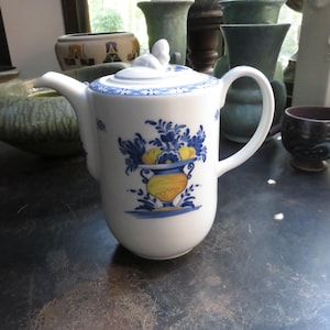 May include: White ceramic teapot with a blue and yellow floral design. The teapot has a curved spout, a large handle, and a lid with a small decorative figure. The design features a vase with fruit and flowers.