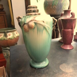 May include: A collection of ceramic vases in various shapes and sizes. The central vase is a tall, green and cream-colored piece with a flared base and handles. Other vases include a maroon vase and a floral-decorated pot.