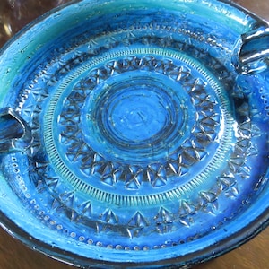 May include: A round, ceramic ashtray with a vibrant blue and turquoise color scheme. The ashtray features a textured surface with concentric circular patterns and three built-in rests for cigarettes. The design includes geometric and dotted details.
