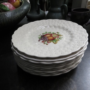 May include: A stack of white ceramic plates with a scalloped edge and a textured surface. Each plate features a central fruit design with apples, grapes, and pears. The plates are stacked on top of each other.