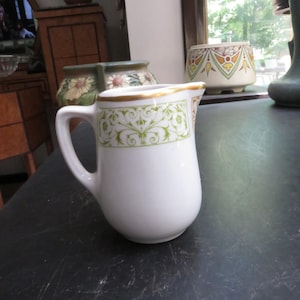 May include: White ceramic creamer with a handle and a decorative green and gold floral pattern around the rim. The creamer is on a dark surface, with other decorative items in the background.