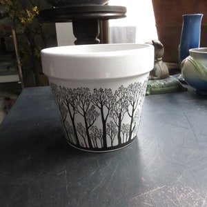 May include: White ceramic planter with a black silhouette design of trees. The planter has a wide rim and a rounded base.
