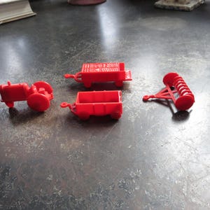 May include: A collection of small, red toy farm vehicles, including a tractor, two wagons, and a plow. The toys are made of plastic and are arranged on a dark, textured surface. The tractor has large red wheels.
