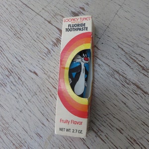 May include: A blue and white cartoon cat on a yellow, orange, and red striped background. The box is for Looney Tunes Anti-Cavity Fluoride Toothpaste, Fruity Flavor, 2.7 oz.