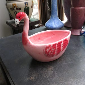 May include: A ceramic bowl shaped like a flamingo with a long, curved neck and a head featuring a red crown, white cheeks, and a black-tipped beak. The body of the bowl is light pink with painted red wing details on the side and a smooth interior.
