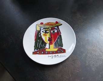 May include: White ceramic plate featuring a colorful abstract portrait in yellow, red, and green, reminiscent of Picasso's style. The plate includes the artist's signature.