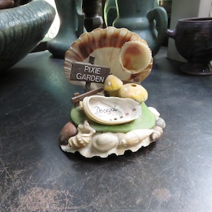 May include: A whimsical fairy garden figurine featuring a large seashell with a smaller shell inside. The base includes a green hill, mushrooms, and a sign that reads "Pixie Garden". The word "Donegal" is written on a shell.
