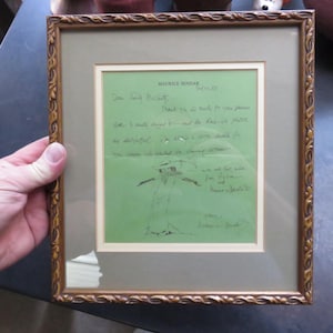 May include: Framed artwork featuring a handwritten letter on green paper, signed by Maurice Sendak. The frame is ornate with a gold finish. The artwork includes a sketch of a figure.