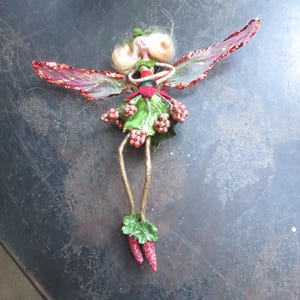 May include: A whimsical fairy figurine with blonde hair, a green dress adorned with red berries, and delicate red and gold glitter wings. The fairy has gold legs and red glitter carrot-shaped feet. This decorative item is perfect for seasonal decor.